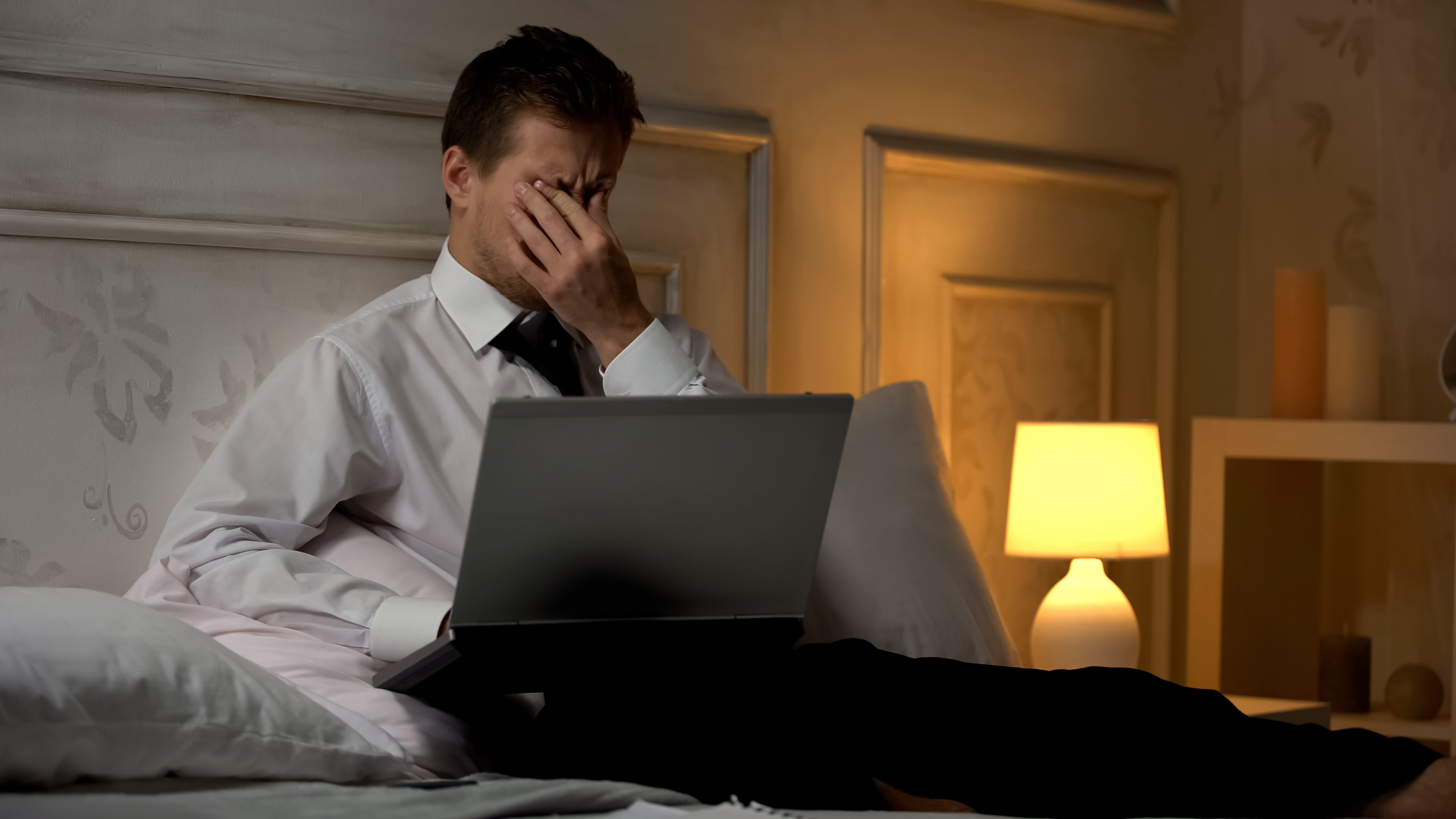 Mand i skjorte og slips sidder i sengen med en bærbar computer og dækker ansigtet med hånden i et dæmpet oplyst rum.