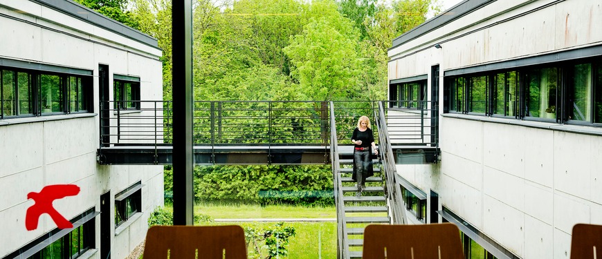 To moderne kontorbygninger forbundet med en gangbro, med en person på en udvendig trappe og grønne træer bagved.