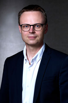 Man wearing glasses, a white shirt, and a navy blazer in a studio portrait.