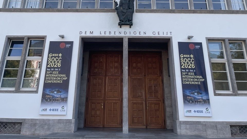 Bygningsindgang med to konferenc bannere for "IEEE SOCC 2026" på hver side af dobbeltdøre.
