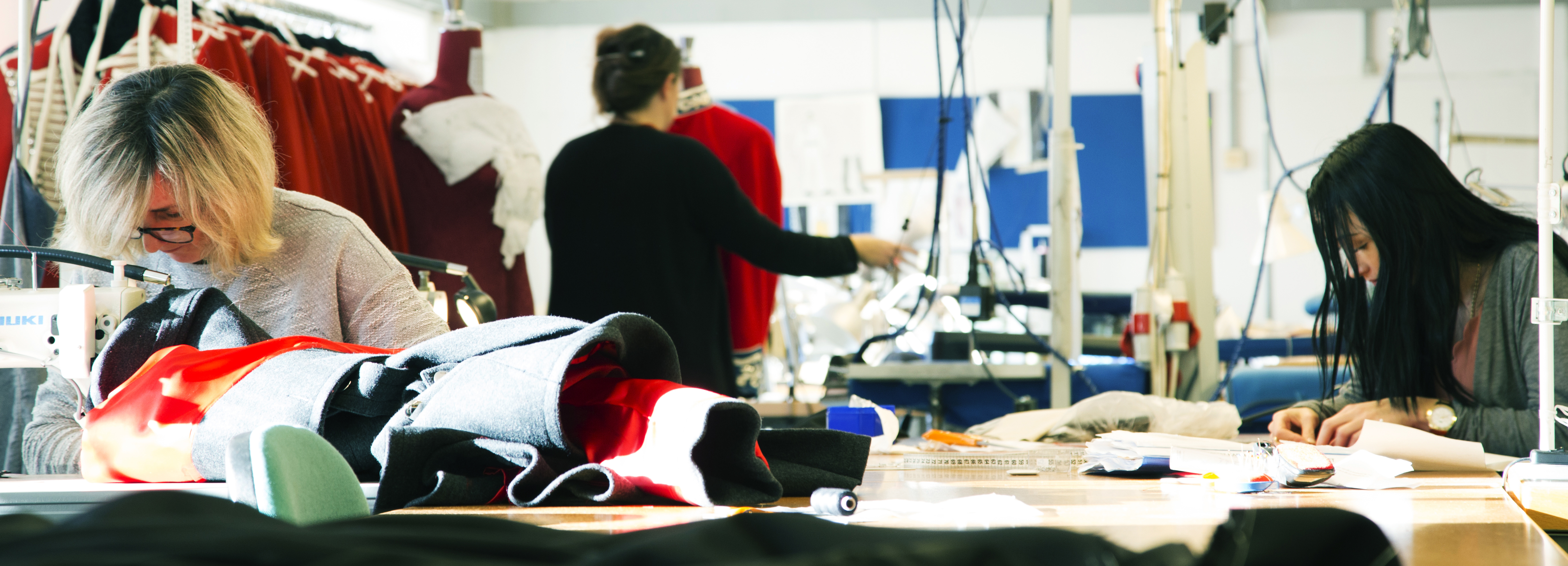 Flere syersker arbejder i et atelier med røde stoffer, mannequin, arbejdsborde og symaskiner.