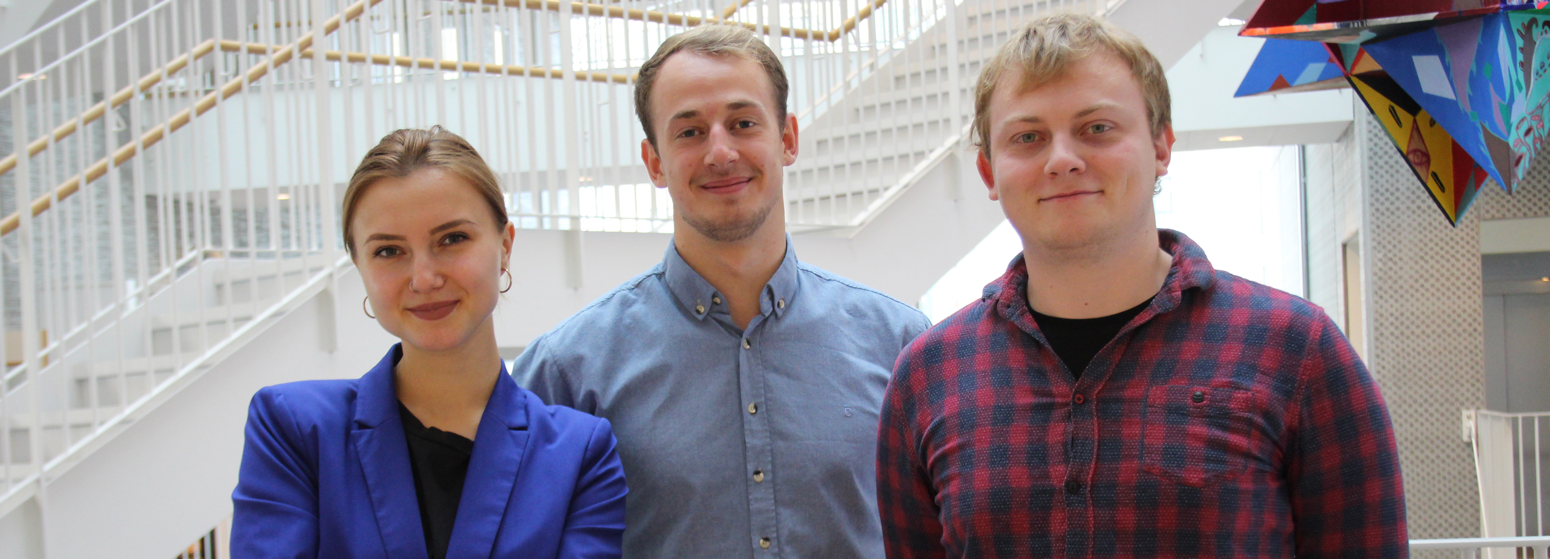 Tre unge personer poserer i et lyst atrium med trapper i baggrunden.