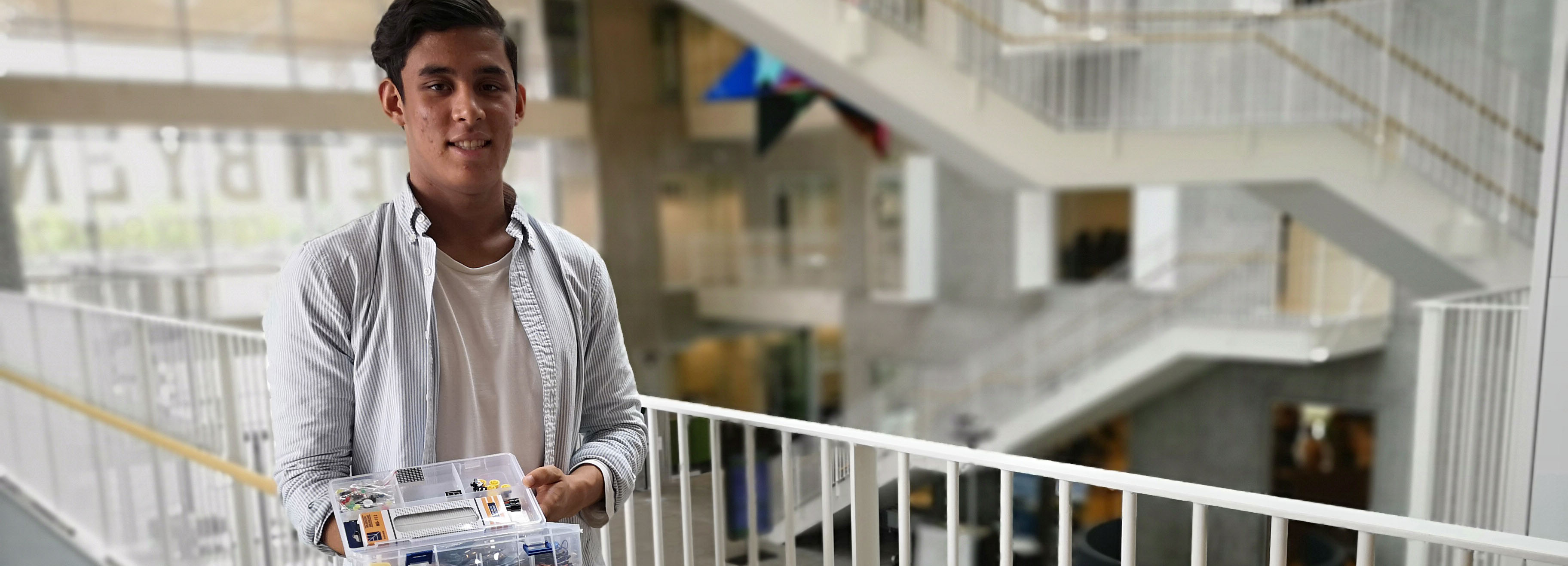 Person, der holder en gennemsigtig kasse med elektronikkomponenter ved et gelænder i en moderne bygning.