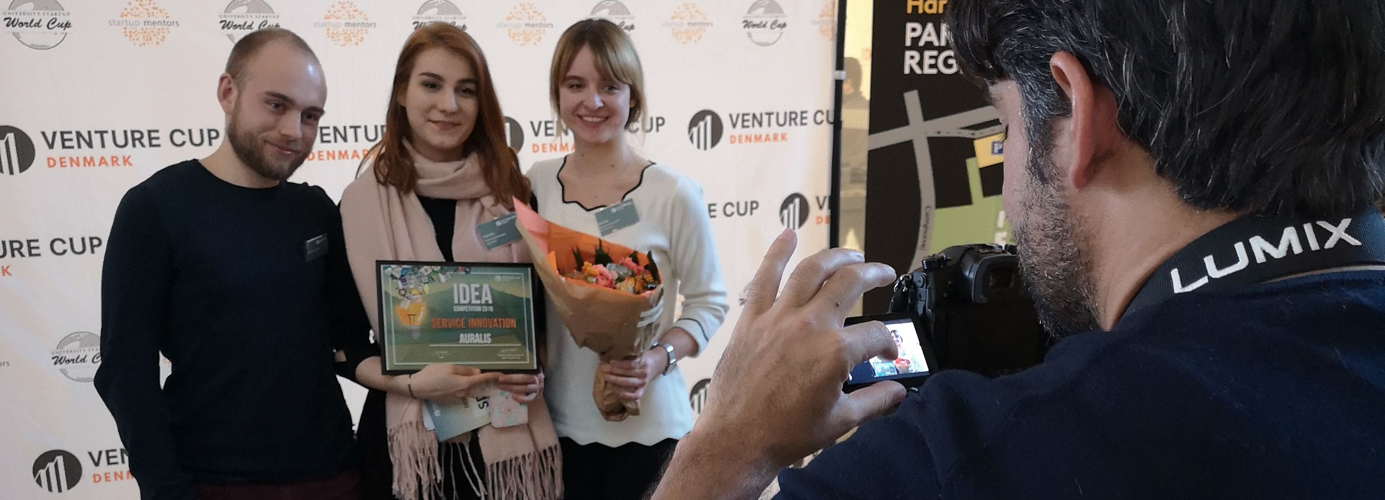 Tre personer fotograferes ved et Venture Cup Denmark-arrangement, hvor de holder et diplom med teksten "IDEA 2016", "SERVICE INNOVATION" og "AURALIS" samt en blomsterbuket.