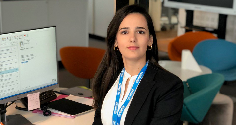 Kvinde i sort blazer ved et skrivebord på et kontor med computerskærm og blåt "UNHCR"-snørrebånd.