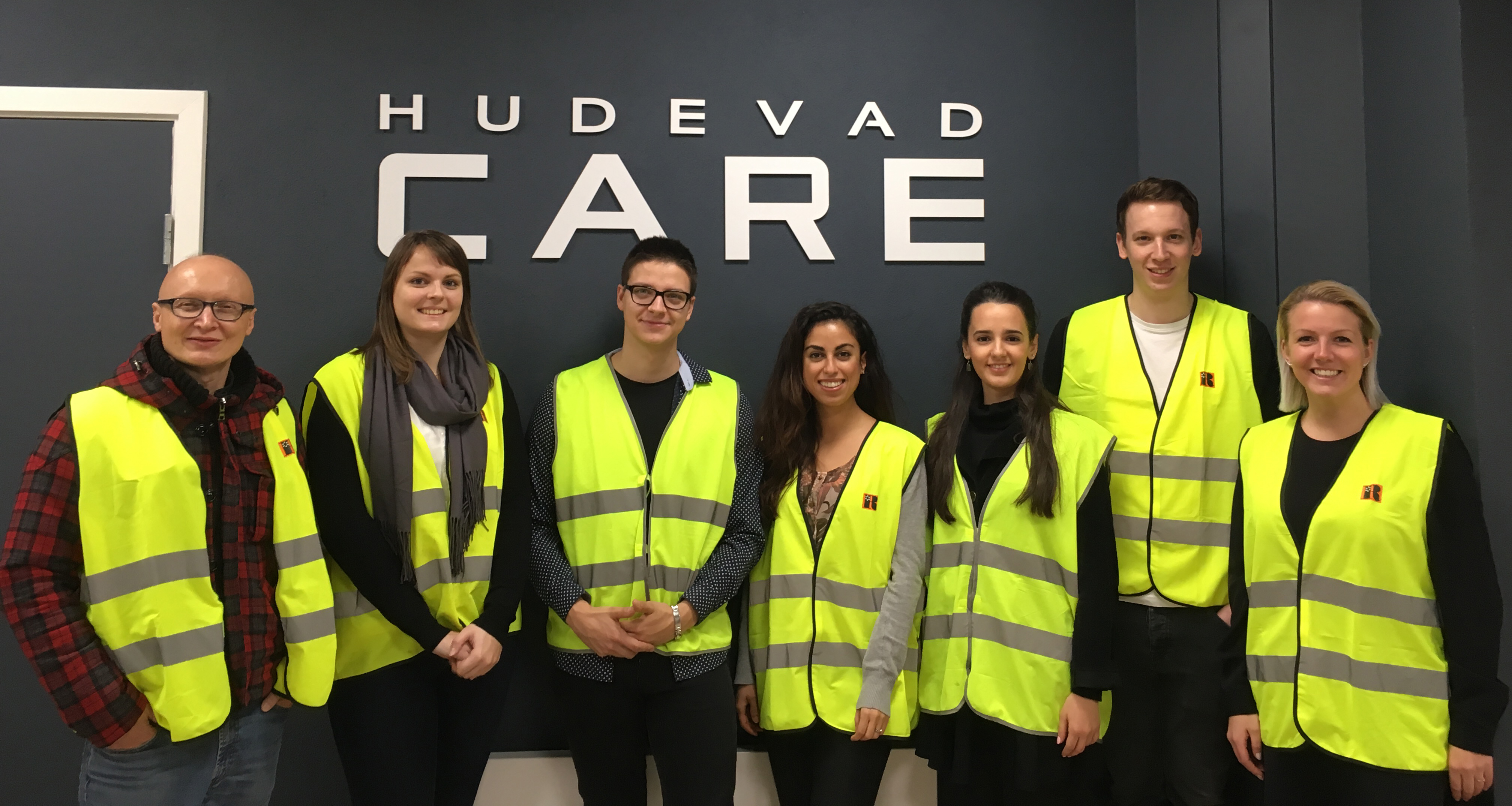 Syv personer i gule sikkerhedsveste står foran skiltet "HUDEVAD CARE".
