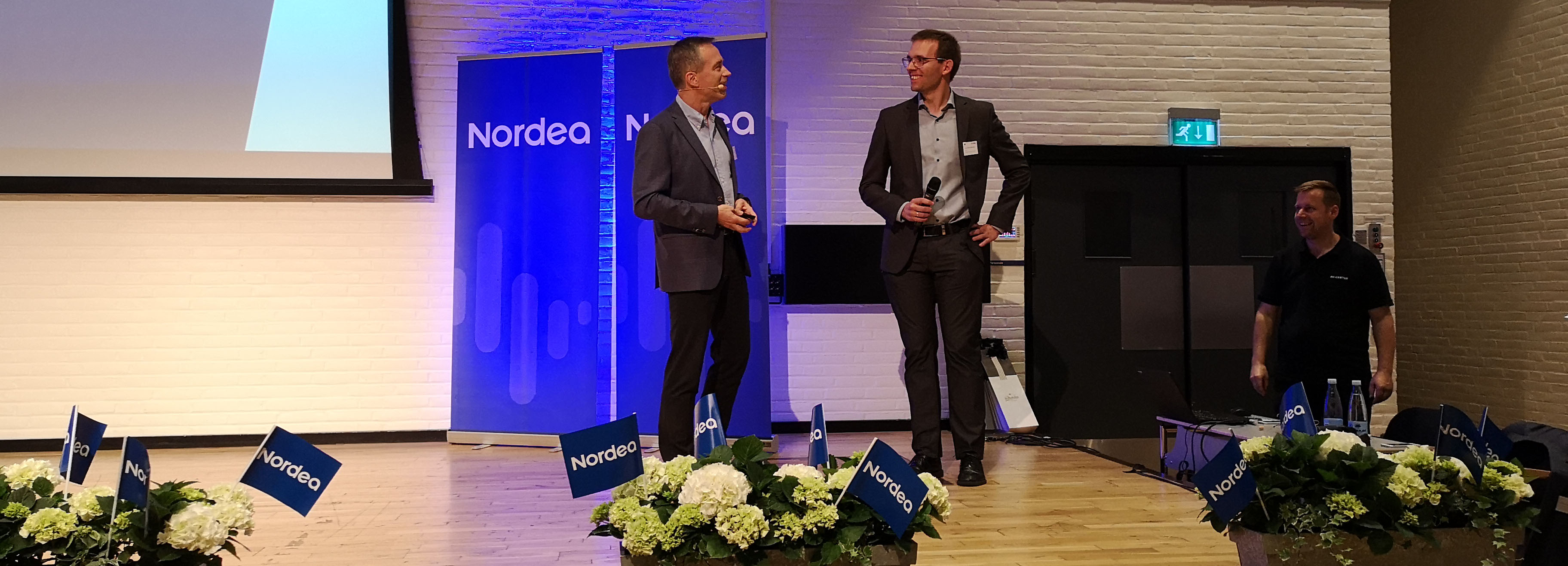 To mænd på en scene ved et arrangement med "Nordea"-bannere, "Nordea"-flag og blomster foran scenen.