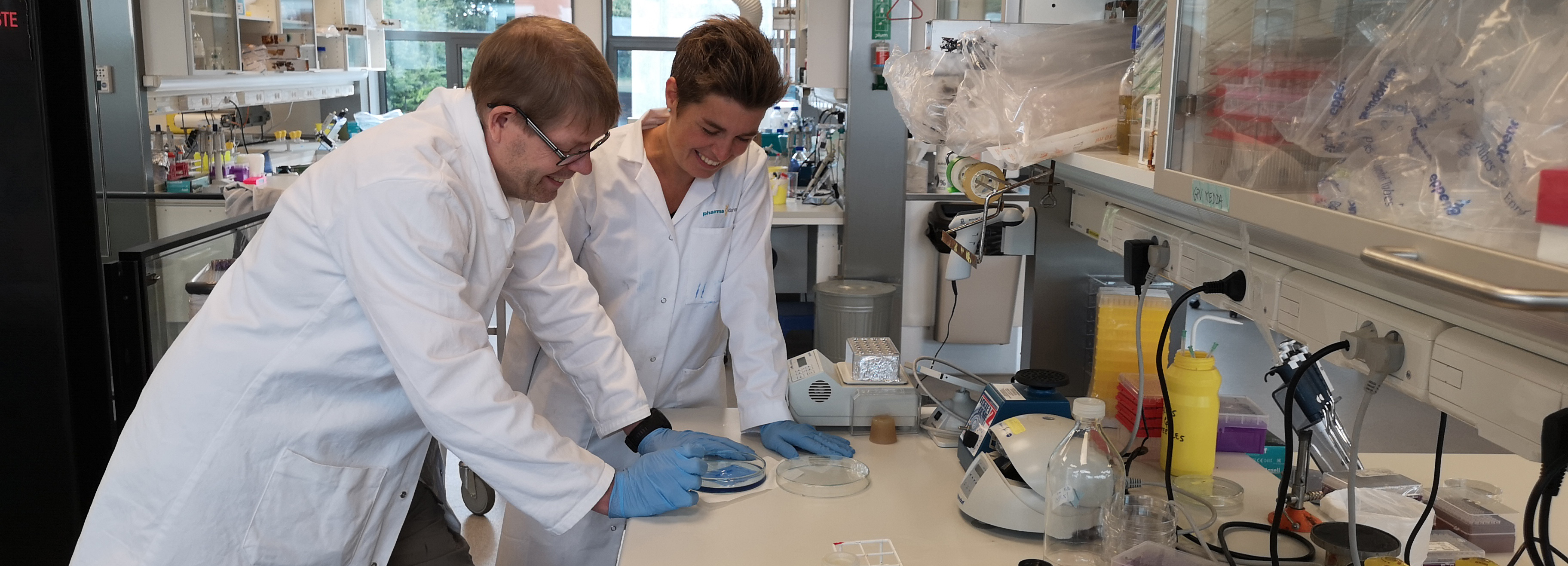To forskere i laboratoriefrakker undersøger petriskåle ved et laboratoriebord omgivet af laboratorieudstyr.