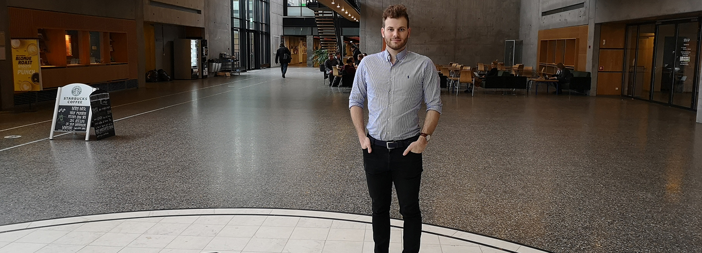 Mand i lys skjorte står i et moderne atrium; til venstre ses et "STARBUCKS COFFEE"-skilt og en plakat med "BLONDE ROAST PUNCH".
