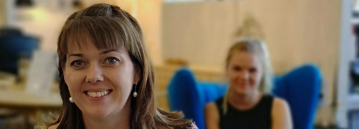 Smilende kvinde i forgrunden med en sløret person i baggrunden indendørs.