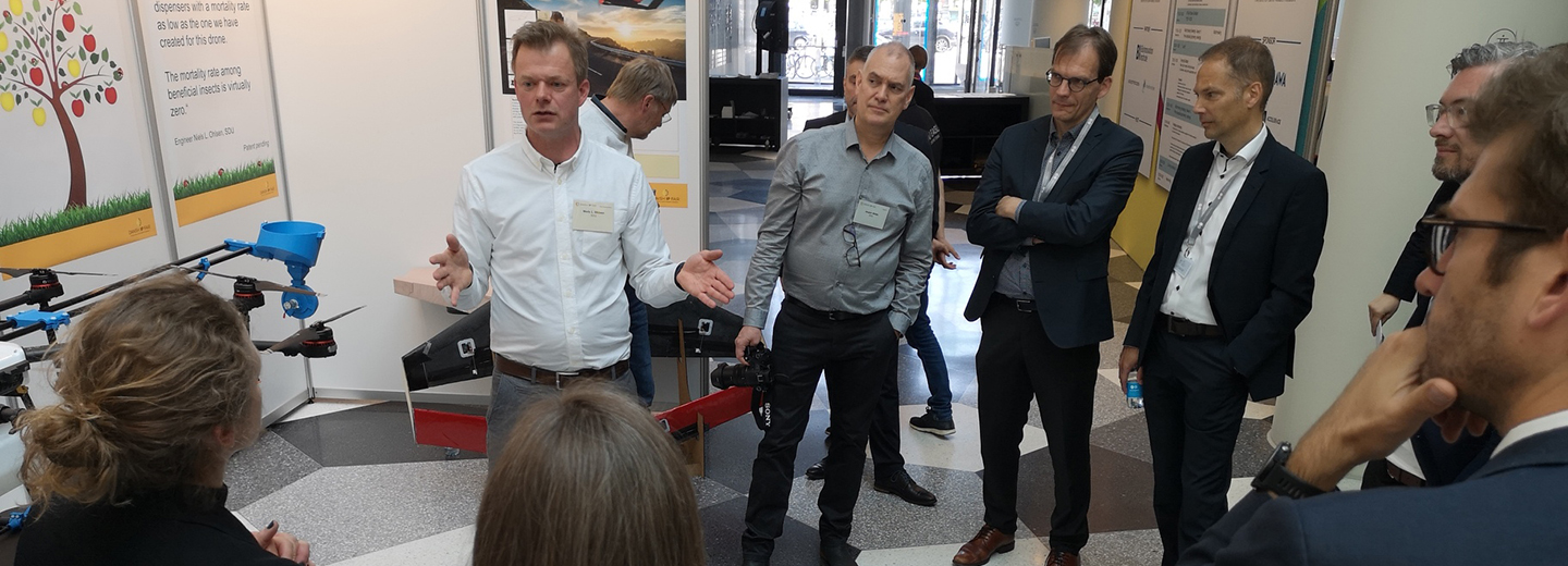 Mand præsenterer en drone med dispenser for en gruppe deltagere ved en indendørs udstilling.