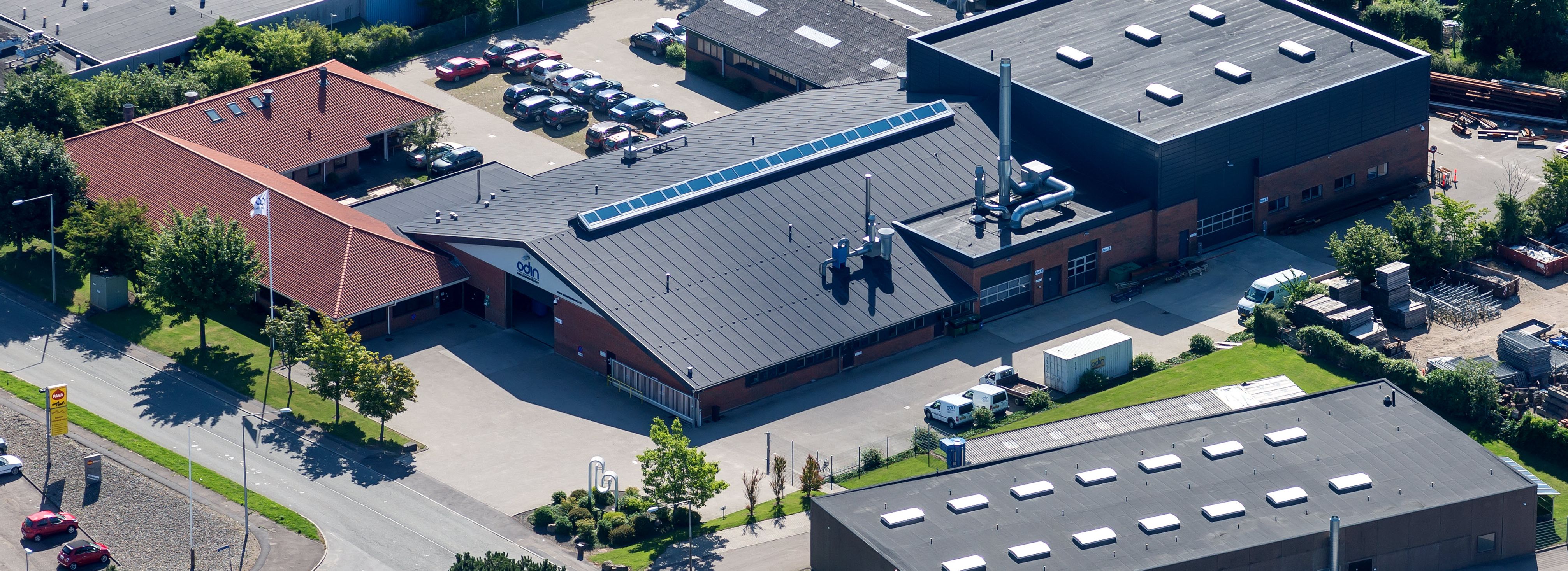 Luftfoto af et industrikompleks med røde og mørke tage, parkeringsplads, varevogne og "odin"-logo på bygninger og udstyr.