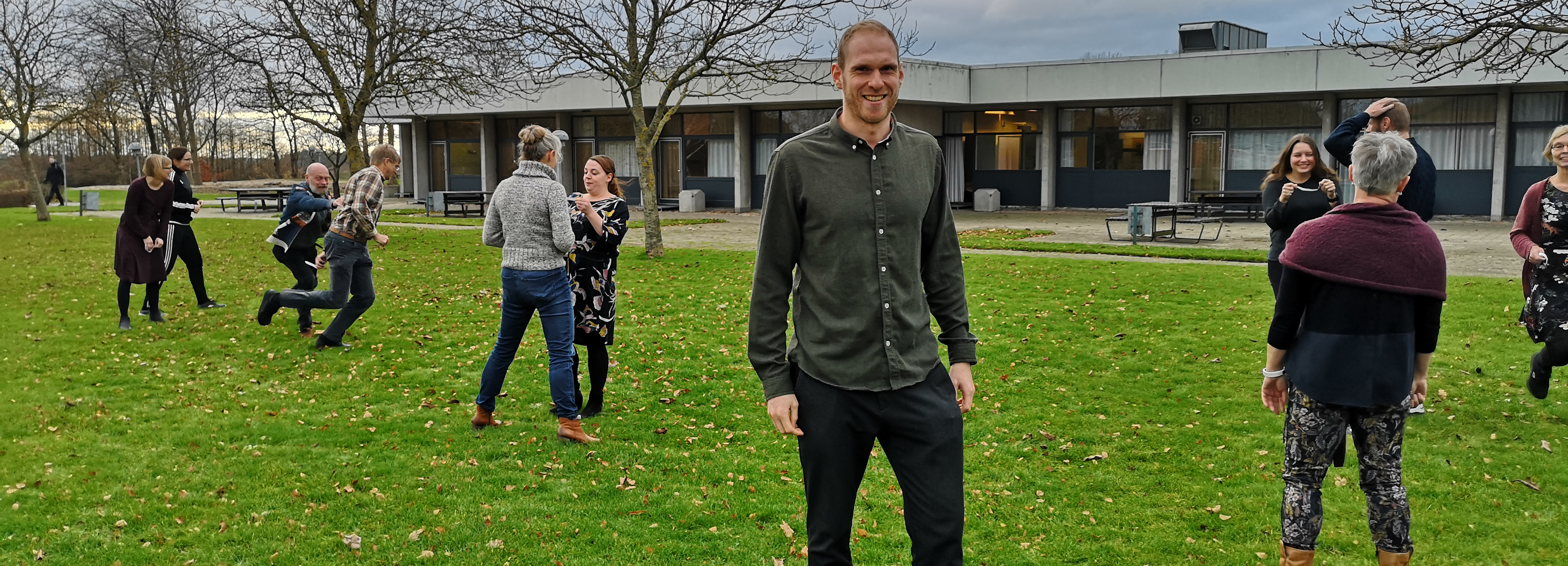 Voksne deltager i en udendørs aktivitet på en græsplæne foran en bygning, med en mand i forgrunden og flere personer i baggrunden.