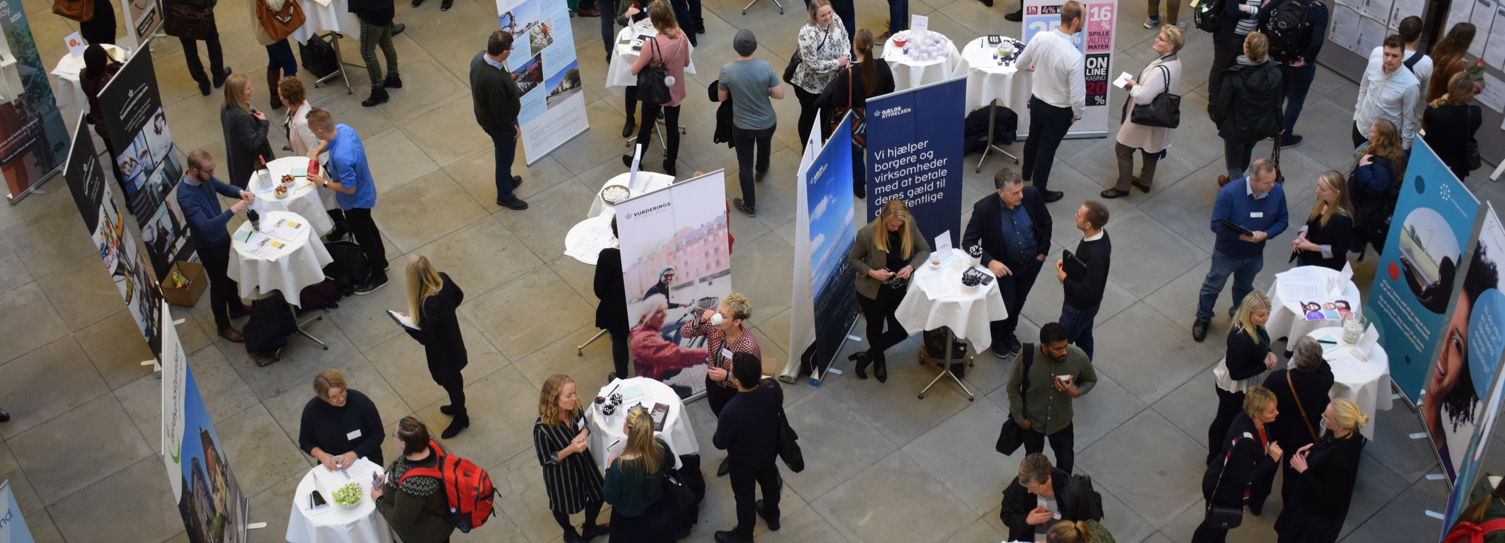 Højt vinkelbillede af en travl indendørs messe med mennesker, ståborde og informationsstande.
