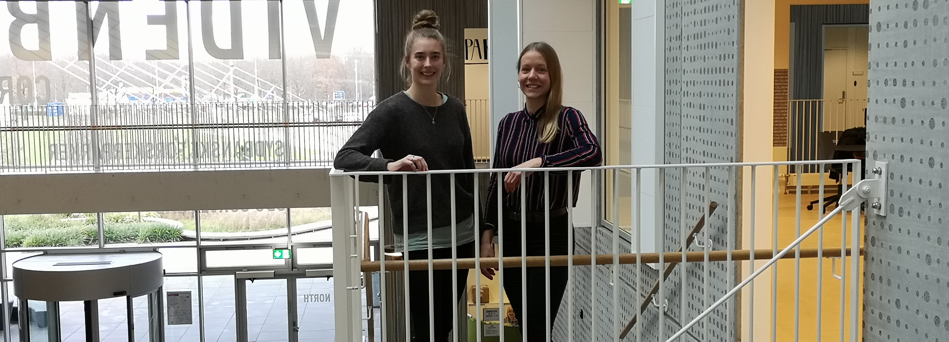 To kvinder står og smiler ved et gelænder i en lys bygning med store vinduer.
