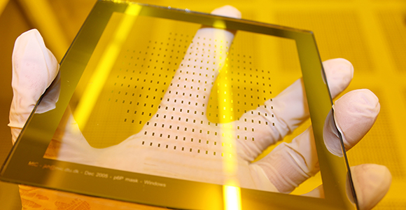 Close-up of a glass wafer with nanostructures held by a hand in a white rubber glove in front of yellow background