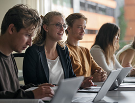 En gruppe SDU studerende, der arbejder ved borde med bærbare computere og papirer i et undervisningsmiljø.