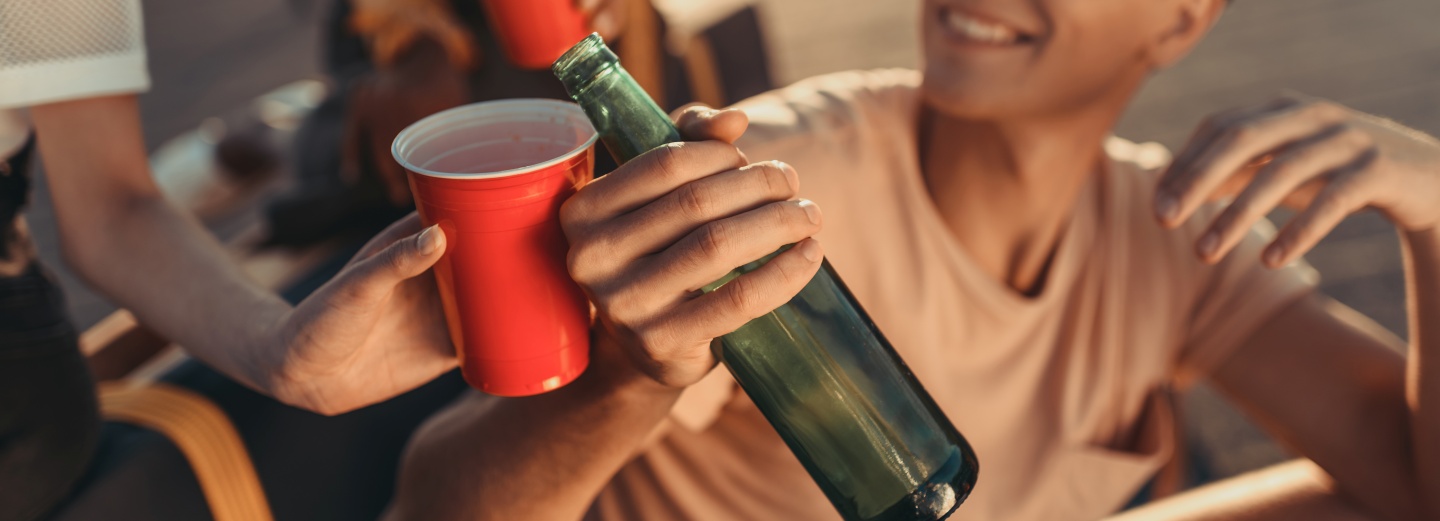 Hænder, der holder en rød plastikkop og en grøn glasflaske under en social sammenkomst udendørs.