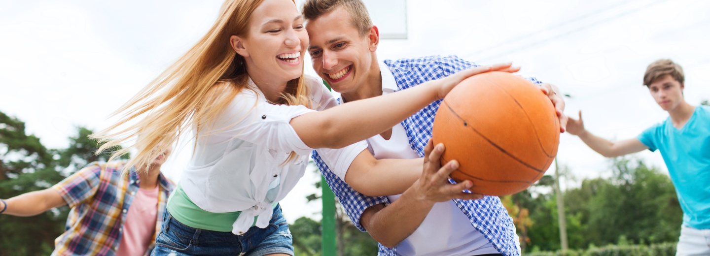 To unge mennesker i forgrunden kæmper om en basketball under et udendørs spil, mens venner ses i baggrunden.