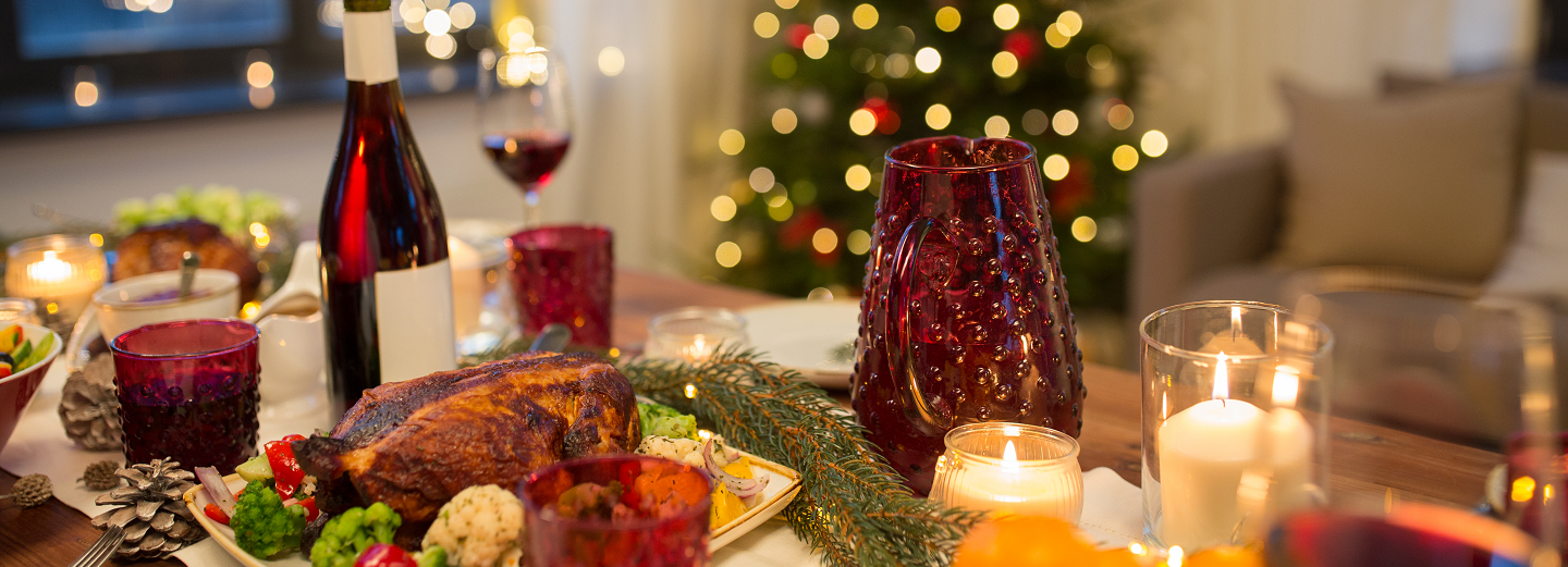 Festligt julebord med helstegt fugl, rødvin, røde glas, levende lys og et oplyst juletræ i baggrunden.