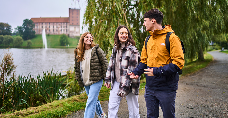 Three SDU students walking along a lakeside path with Koldinghus and fountain in the background.