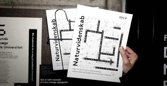 En hånd holder et SDU-magasin om naturvidenskabelige uddannelser foran en opslagstavle med informationsmateriale.