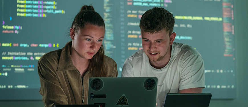 To studerende i undervisningslokale med kode på skærm i baggrunden