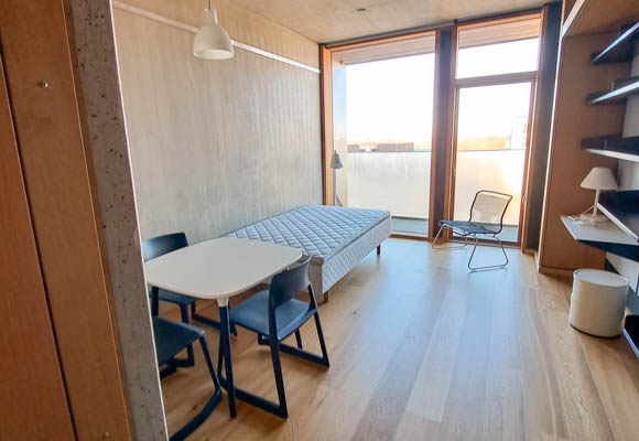 Billedet viser lejlighedens sovesofa op ad venstre væg og hylder på den højre væg. I baggrunden er panoramavindue med udgang til altanen.