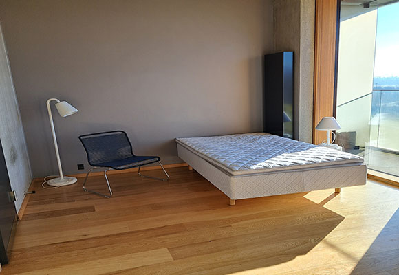 På billedet ses lejlighedens opholdsrum med dør til badeværelset i venstre side, sovesofa med lampe og stol midtfor samt panoramavindue ud til altanen til højre.