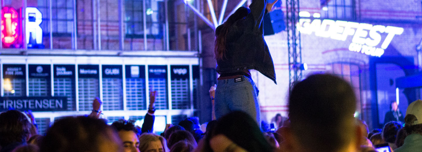 Publikum foran en oplyst facade under en koncert, hvor en person står hævet over mængden med armene oppe.