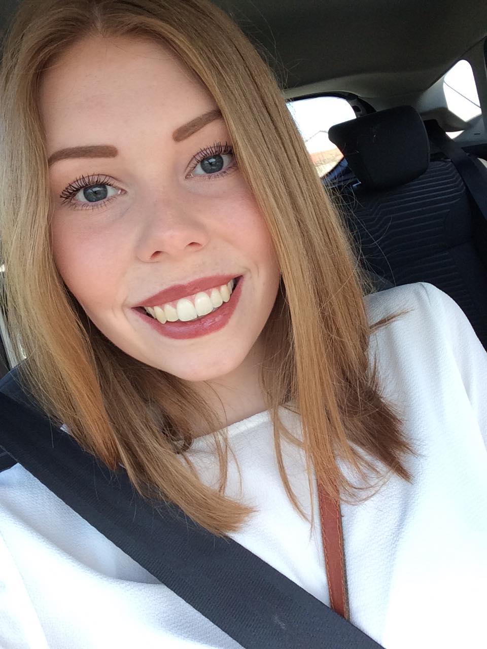 Smilende kvinde med lyst hår tager en selfie i en bil med sikkerhedssele på.