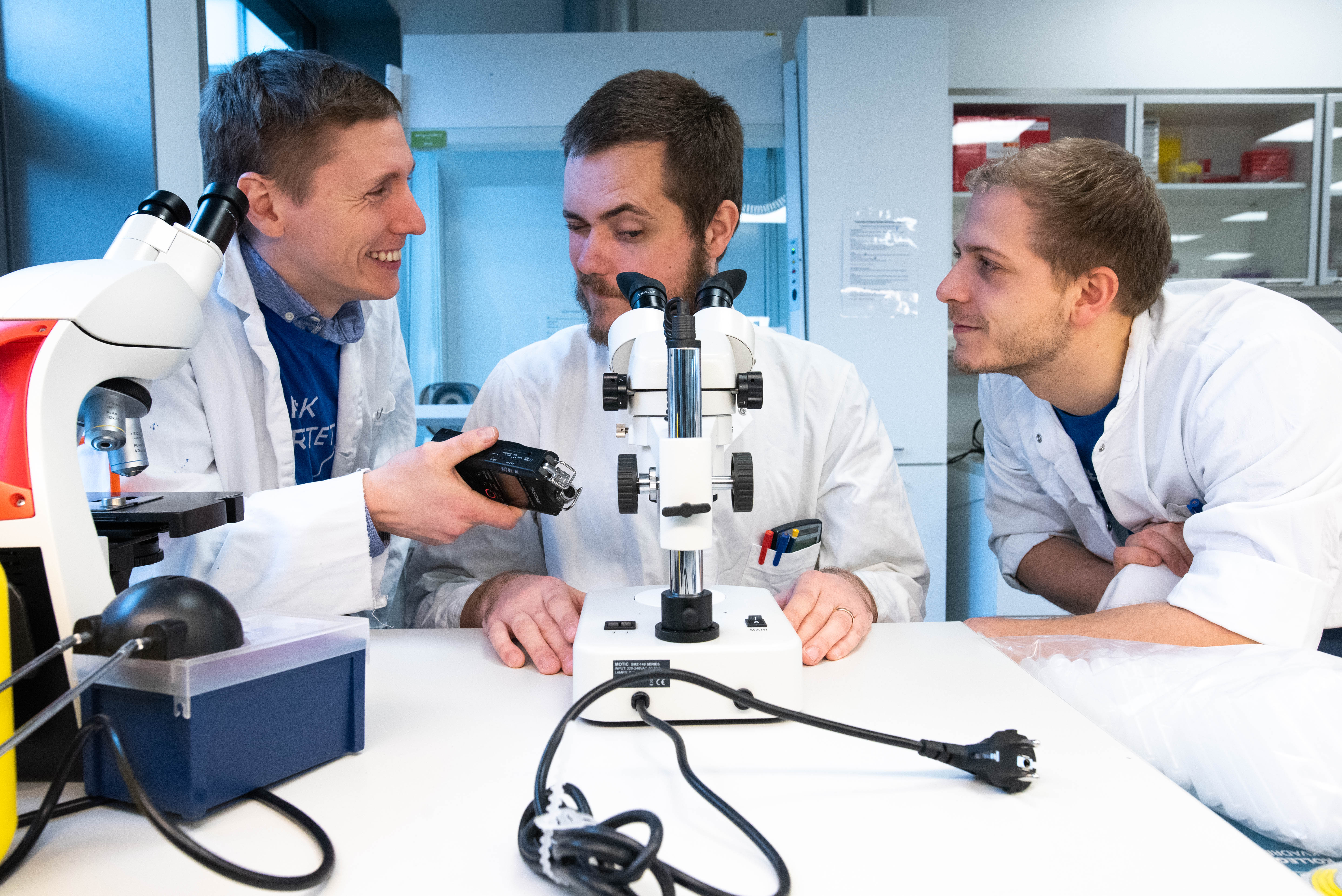 Tre forskere i hvide kitler ved et bord med mikroskoper og laboratorieudstyr