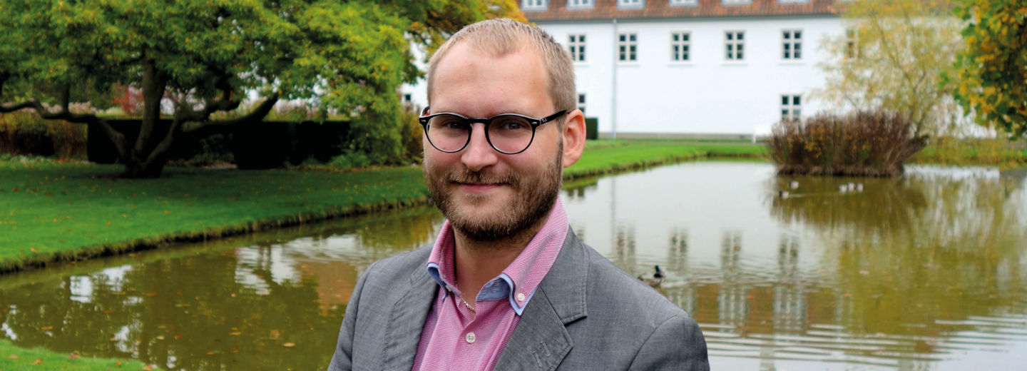 Portræt af en mand med briller og skæg foran en sø og en hvid bygning.