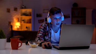 Ung mand med hovedtelefoner bruger en bærbar computer ved et bord i et dæmpet oplyst rum om aftenen.