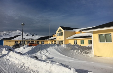 Gul bygning med mørkt tag omgivet af store snedriver og snedækkede bjerge i baggrunden.