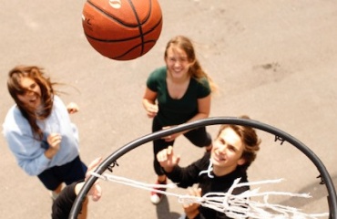 Tre personer spiller basketball på en asfaltbane set oppefra med en bold over kurven.