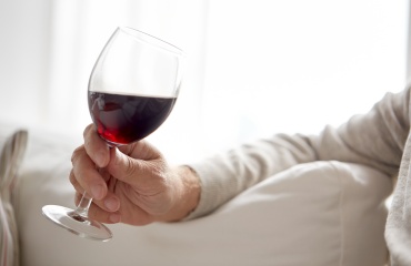 Hånd holder et glas rødvin foran en lys sofa.