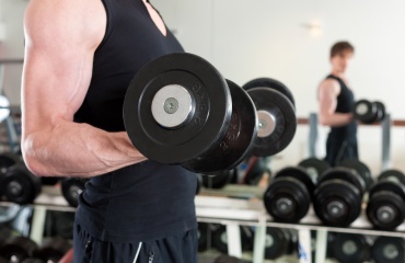Mand løfter en håndvægt under bicepstræning i et fitnesscenter med spejl og vægte i baggrunden.