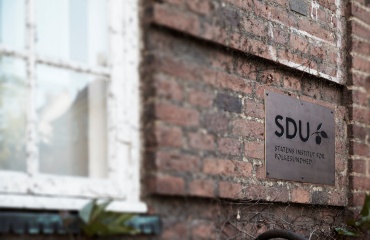 Skilt på murstensvæg med teksten "SDU" og "STATENS INSTITUT FOR FOLKESUNDHED" ved siden af et vindue.