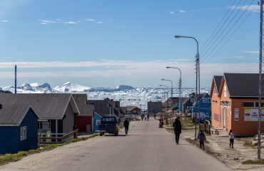 Grusvej i grønlandsk boligområde med farvede huse, mennesker og isbjerge i baggrunden.