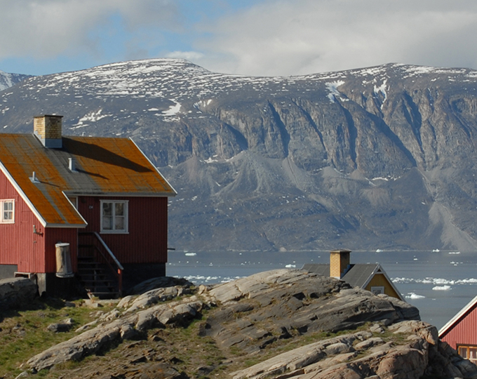 Rødt hus på klipper ved kysten med havis og sneklædt fjeld i baggrunden.