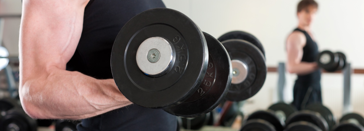 Person løfter sorte håndvægte i et fitnesscenter med en anden person i baggrunden.